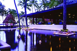A serene pool area with a thatched-roof structure, palm trees, and a covered seating area.