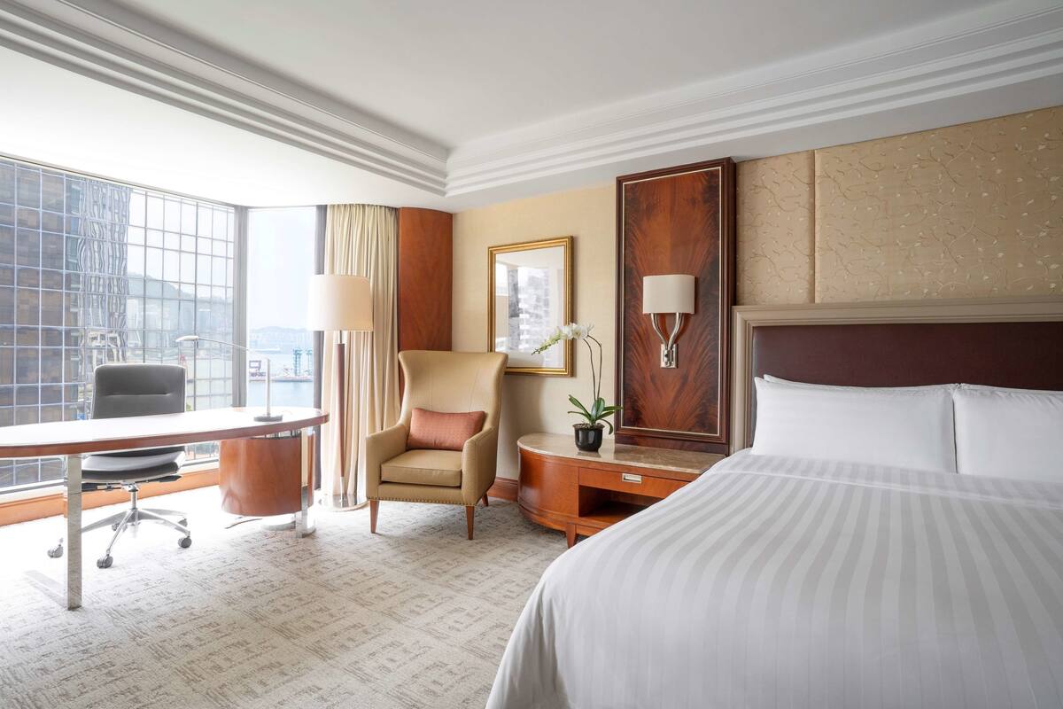 A hotel room with a large bed, a desk with a chair, a small table with a lamp, a potted plant, and a large window with curtains.