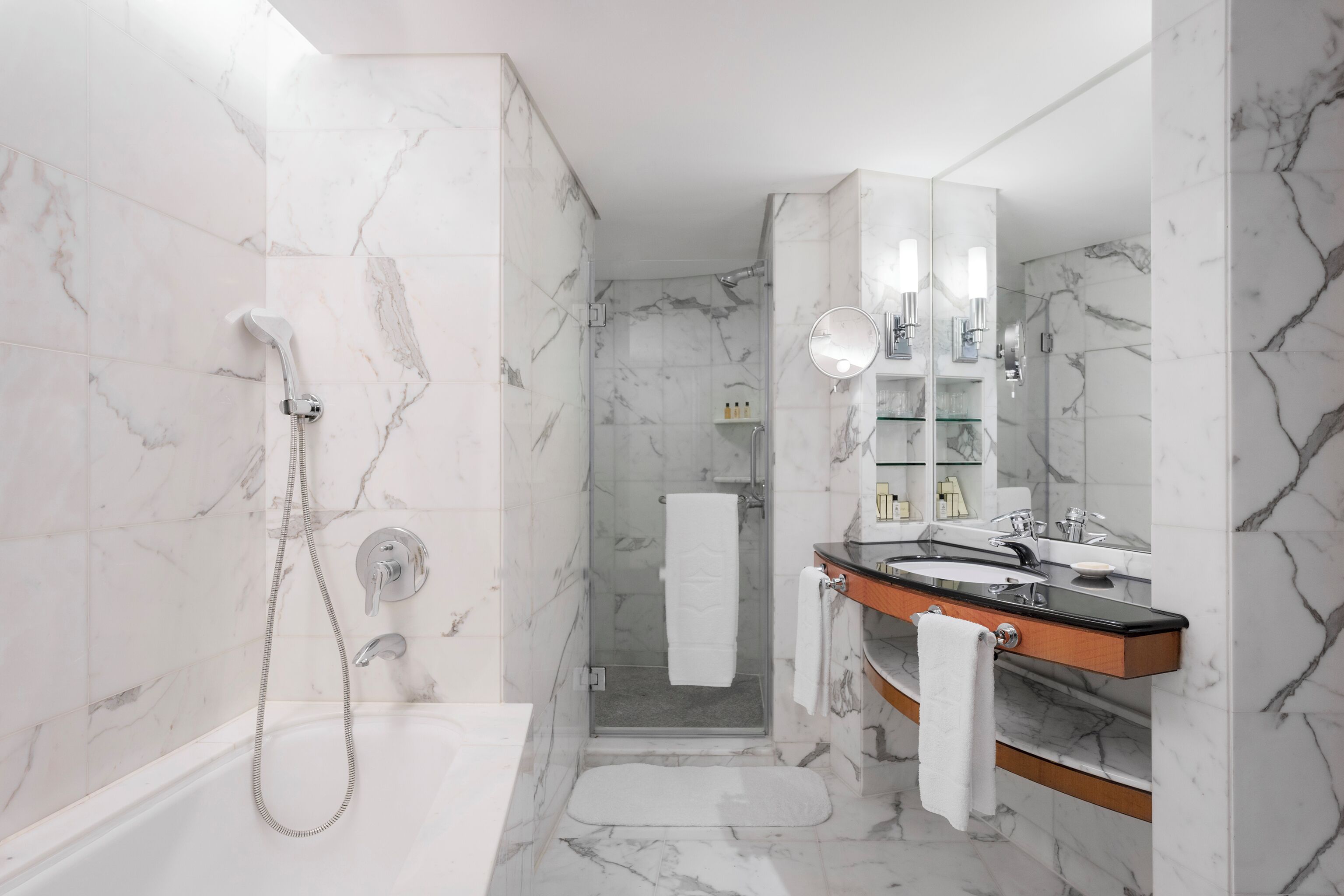 Un baño moderno con ducha envuelta en vidrio, suelo de baldosas de mármol, tocador de madera con encimera negra y un espejo grande.