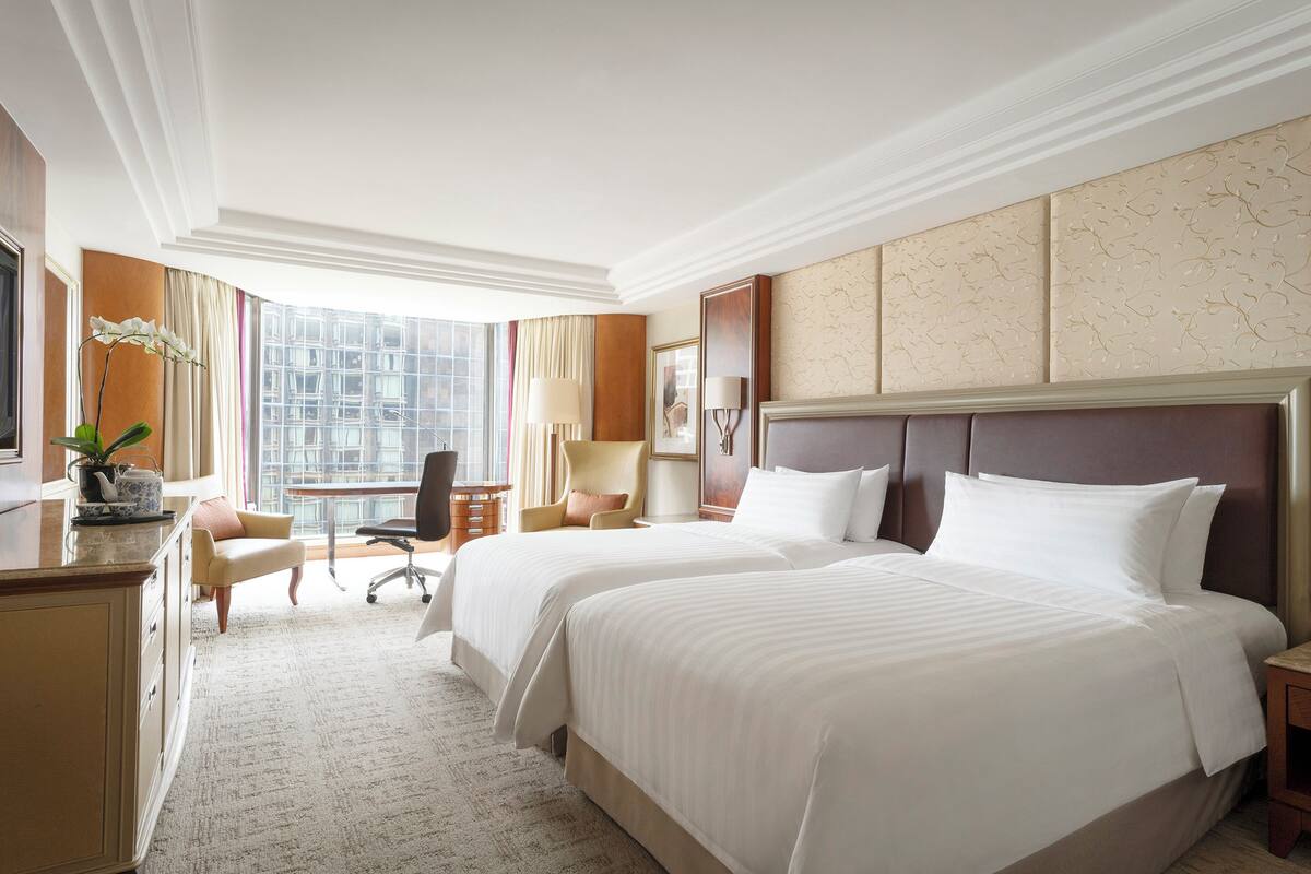 A hotel room with two beds, a desk, a chair, a lamp, and a view of a cityscape.