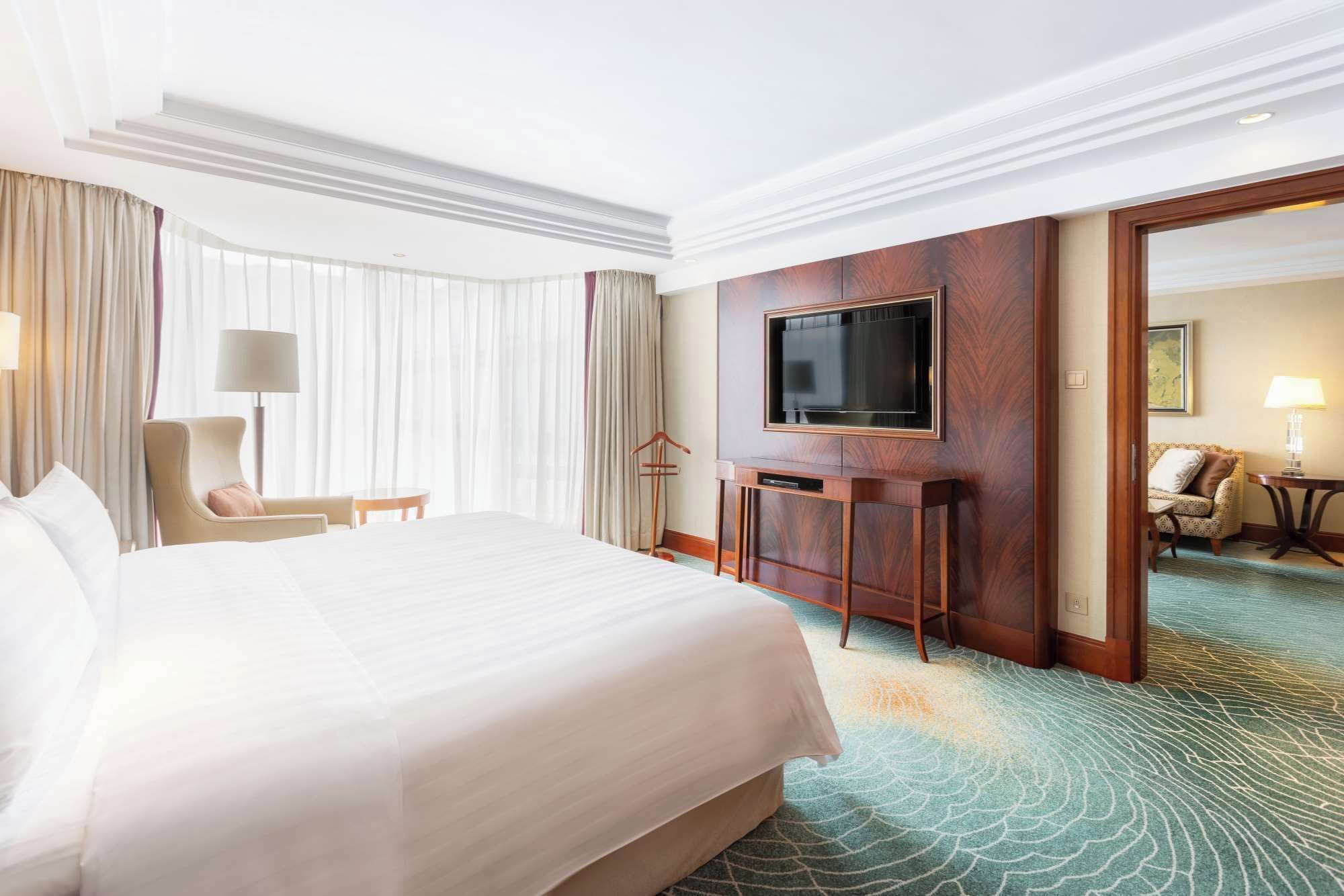 Ein Hotelzimmer mit einem großen Bett, einem an einer Holzplatte montierten Fernseher, einem Schreibtisch mit Lampe und Blick auf ein weiteres Zimmer durch eine offene Tür.