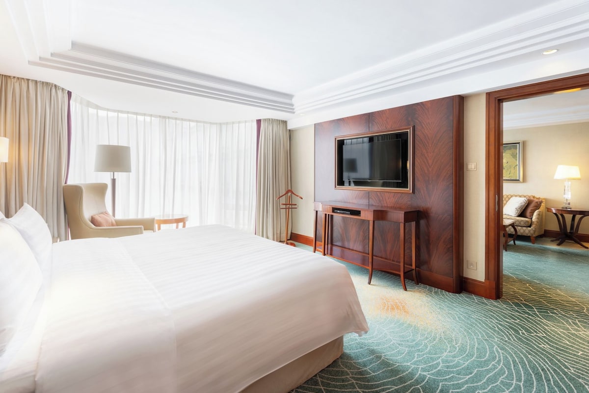 A hotel room with a large bed, a flat-screen TV mounted on a wooden panel, a seating area with a chair and a small table, and a view through an open door.
