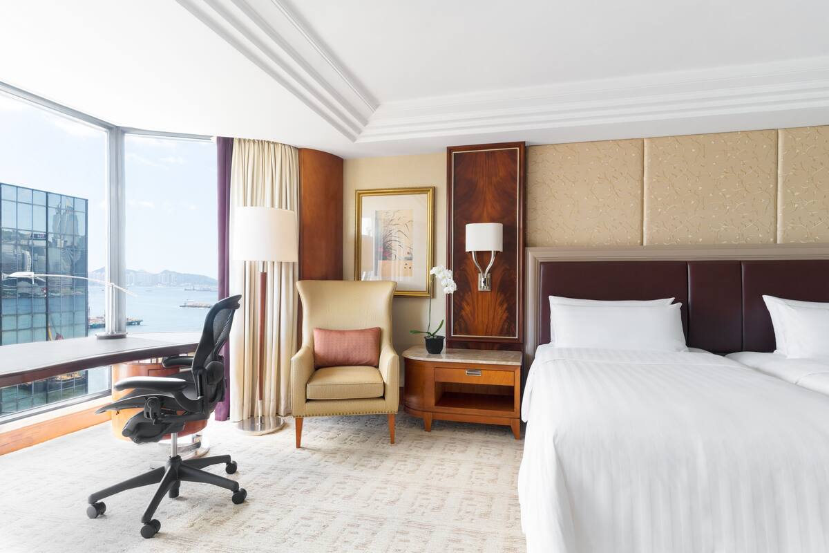 A hotel room with a large bed, a desk with a chair, a nightstand, a lamp, a potted plant, and a view of a cityscape through large windows.