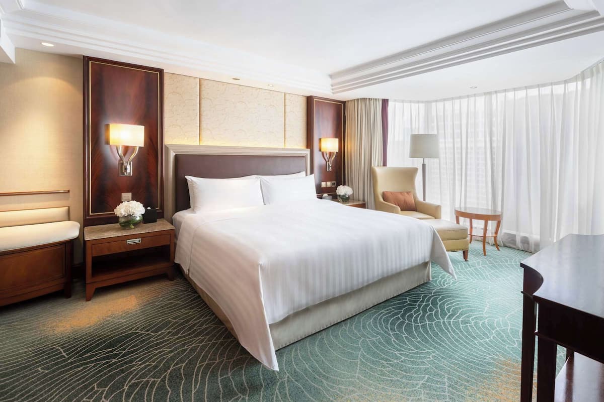 A hotel room with a large bed, two bedside tables, a chair, a small table, and a view of the outdoors through sheer curtains.