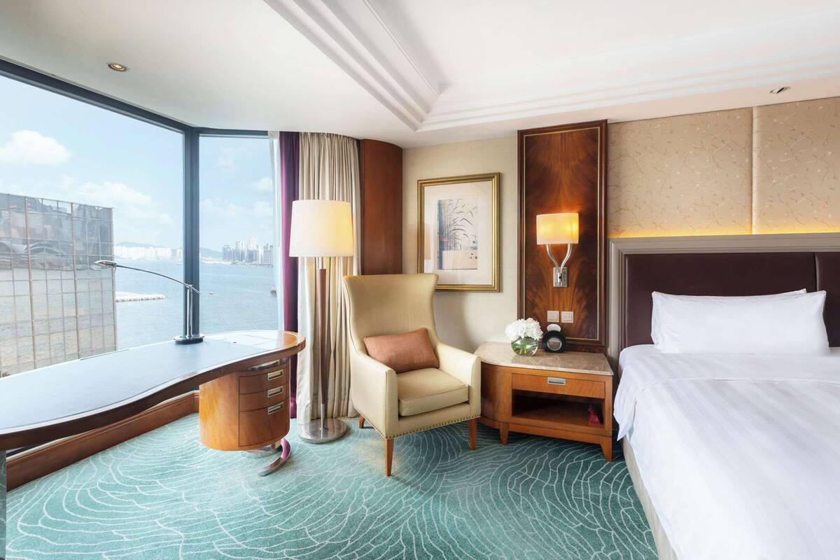 A hotel room with a large window offering a city view, a bed, a chair, a desk, and a nightstand with a lamp.