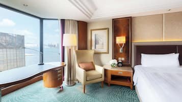 Hotelkamer met groot raam en uitzicht op de stad, een bed, fauteuil, bureau en nachtkastje met lamp.