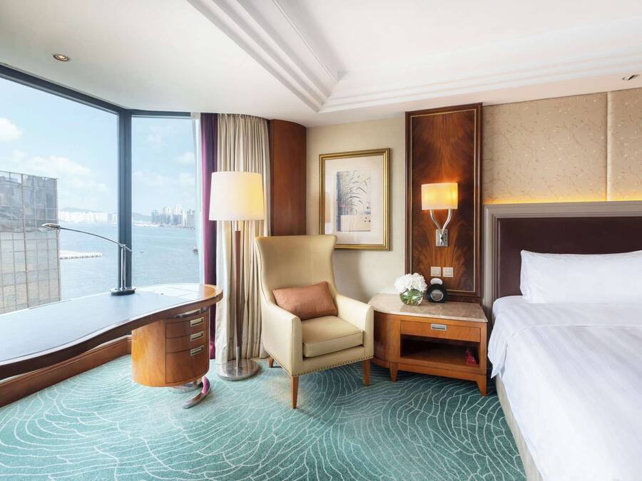 Hotelkamer met groot raam en uitzicht op de stad, een bed, fauteuil, bureau en nachtkastje met lamp.