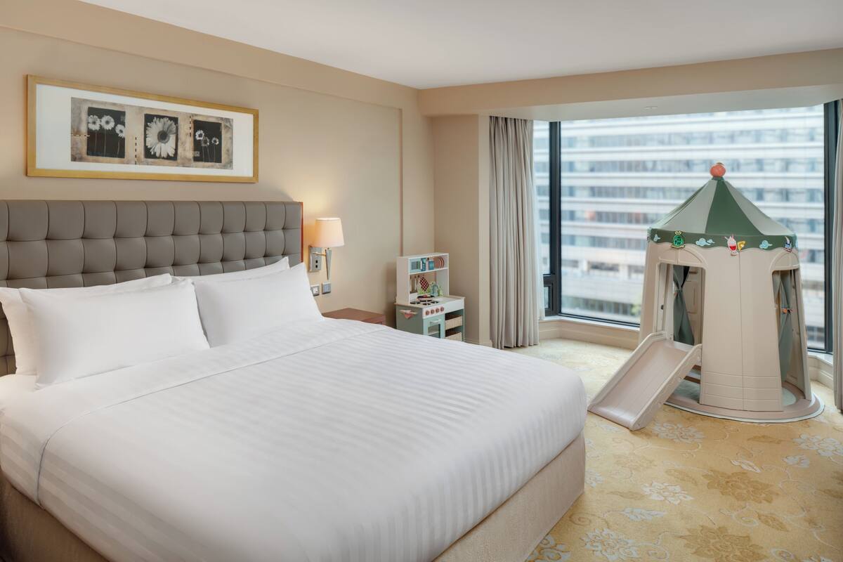 A hotel room with a large bed, a nightstand with a lamp, a bookshelf, a window with curtains, and a children's play area.