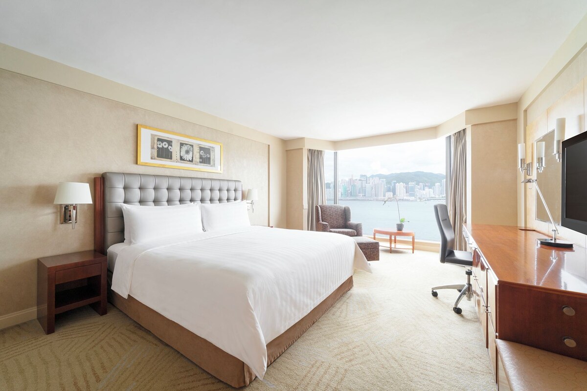 A hotel room with a large bed, a desk, a chair, a TV, and a view of a cityscape.