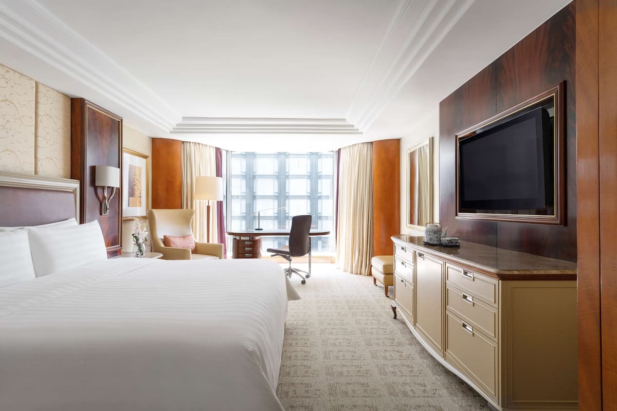 A hotel room with a large bed, a desk with a chair, a television, and a view of the city through large windows.