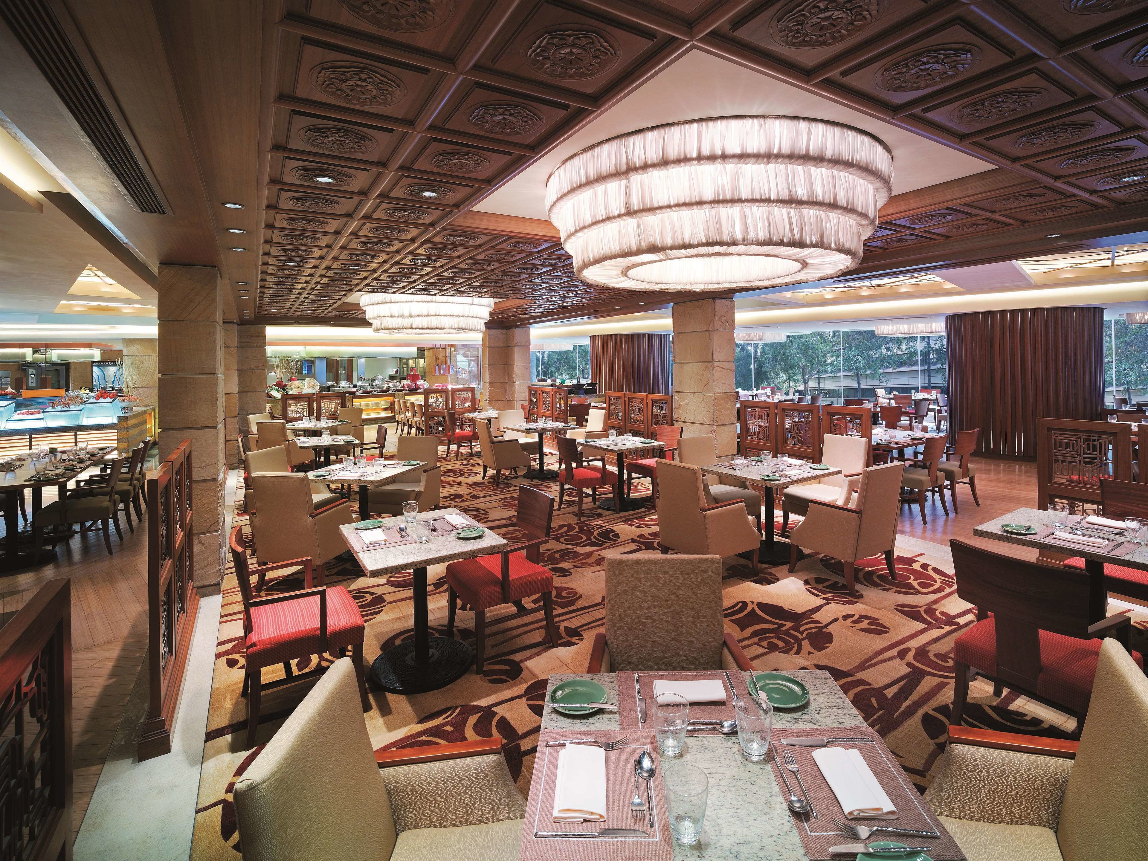 A spacious restaurant with multiple dining tables and chairs, a large chandelier, and a view of the outdoors through large windows.