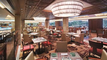 A spacious restaurant with multiple dining tables and chairs, a large chandelier, and a view of the outdoors through large windows.