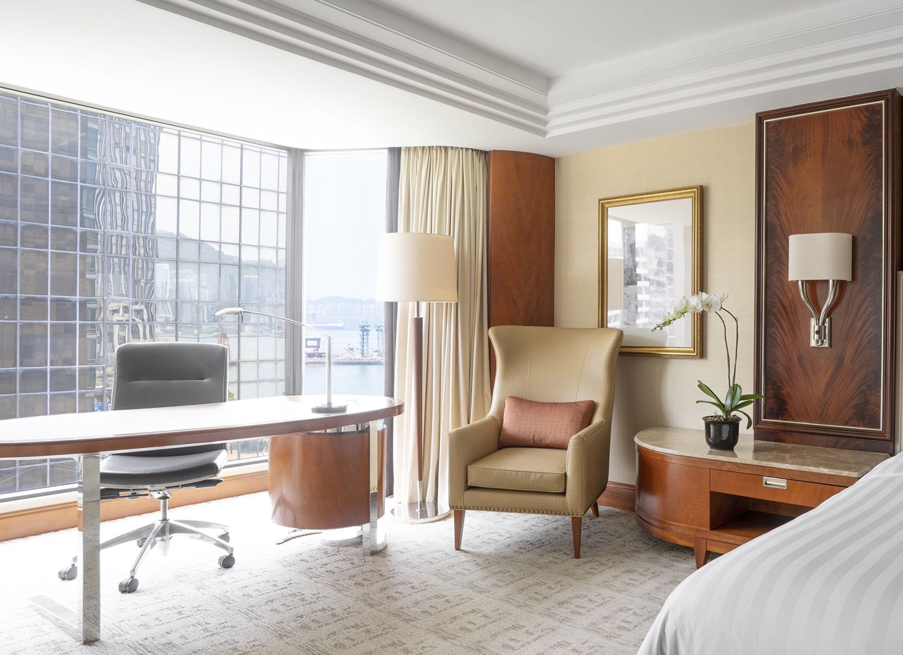 A hotel room with a large window, a desk, a chair, a bedside table, a lamp, and a potted plant.