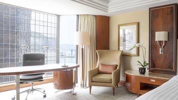 A hotel room with a large window, a desk, a chair, a bedside table, a lamp, and a potted plant.