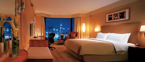 A hotel room with a large bed, a desk, a chair, a nightstand, and a view of a cityscape at night.