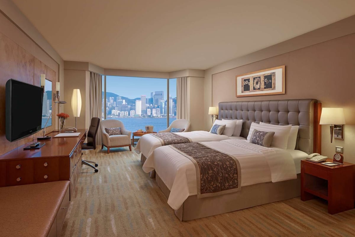 A hotel room with a large bed, two bedside tables, a desk with a TV, a chair, and a view of a city skyline.