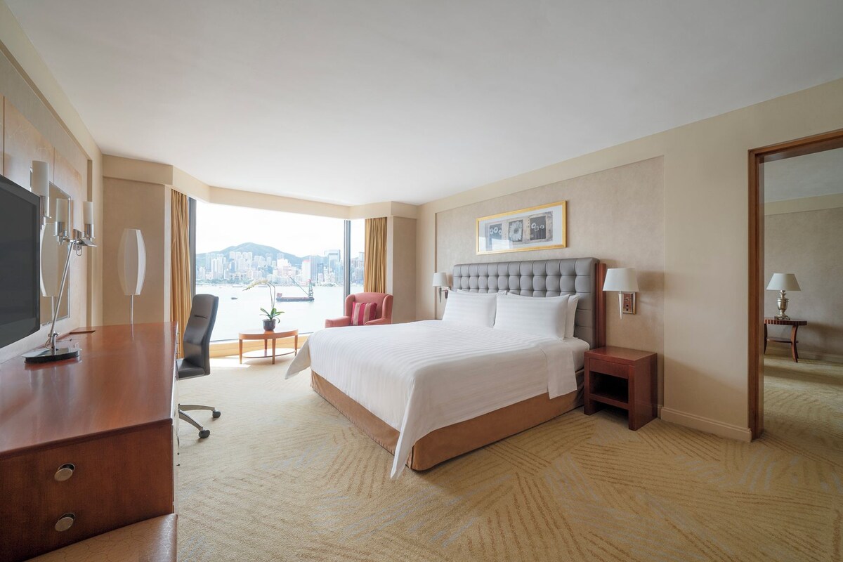 A hotel room with a large bed, a desk with a TV, a chair, a small table with a plant, and a view of a cityscape.