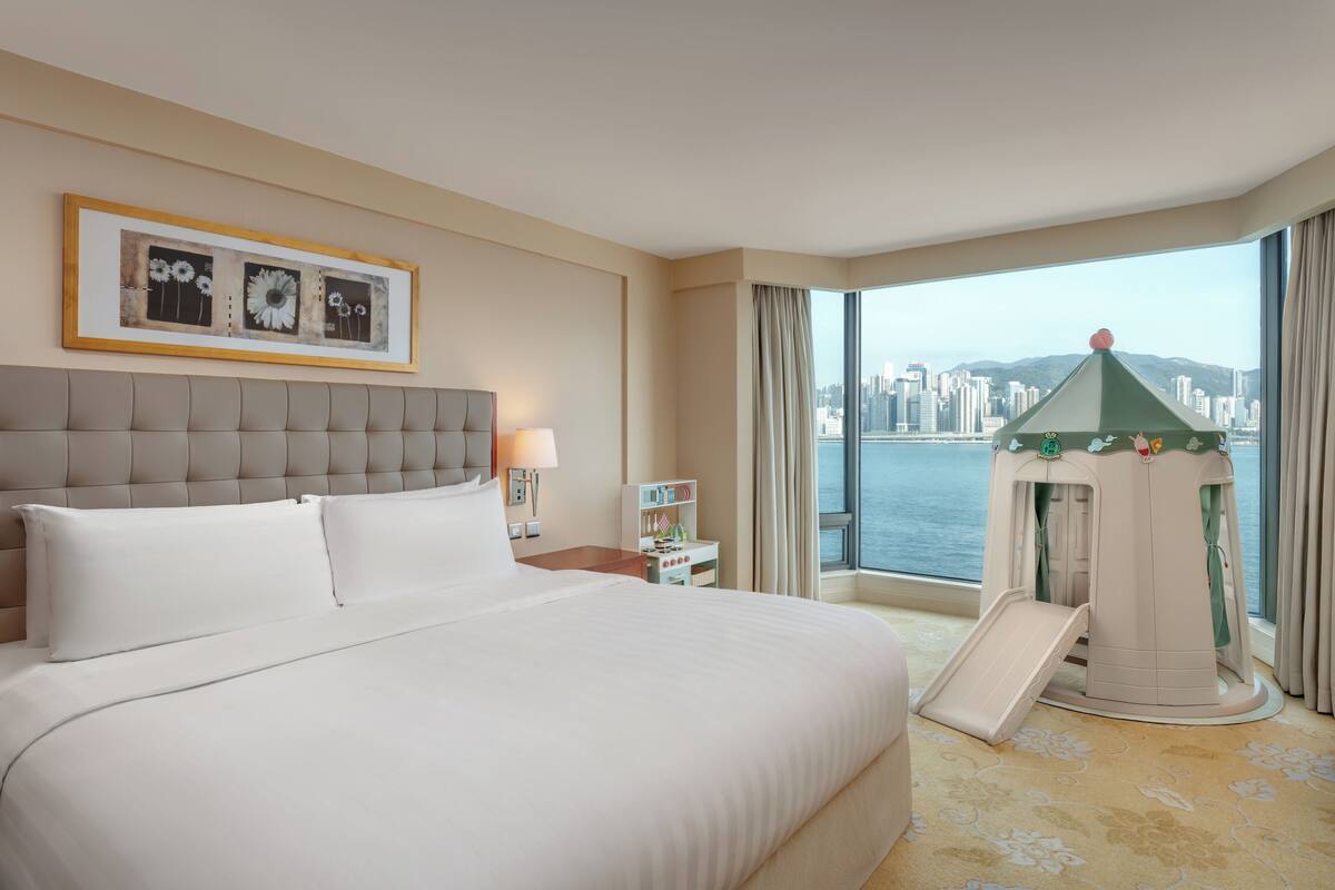 A hotel room with a large bed, a city view, a children's play area, and a framed artwork on the wall.