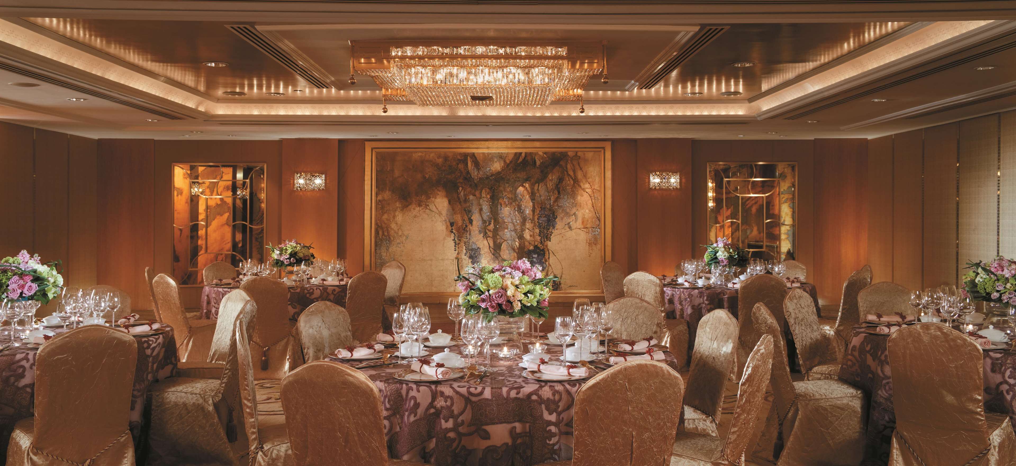 A banquet hall with round tables set for dining, large chandeliers, and a large abstract painting on the wall.