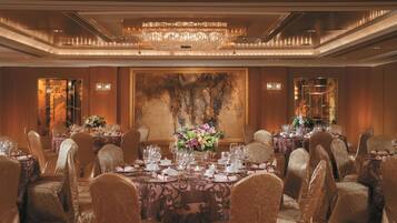 A banquet hall with round tables set for dining, large chandeliers, and a large abstract painting on the wall.