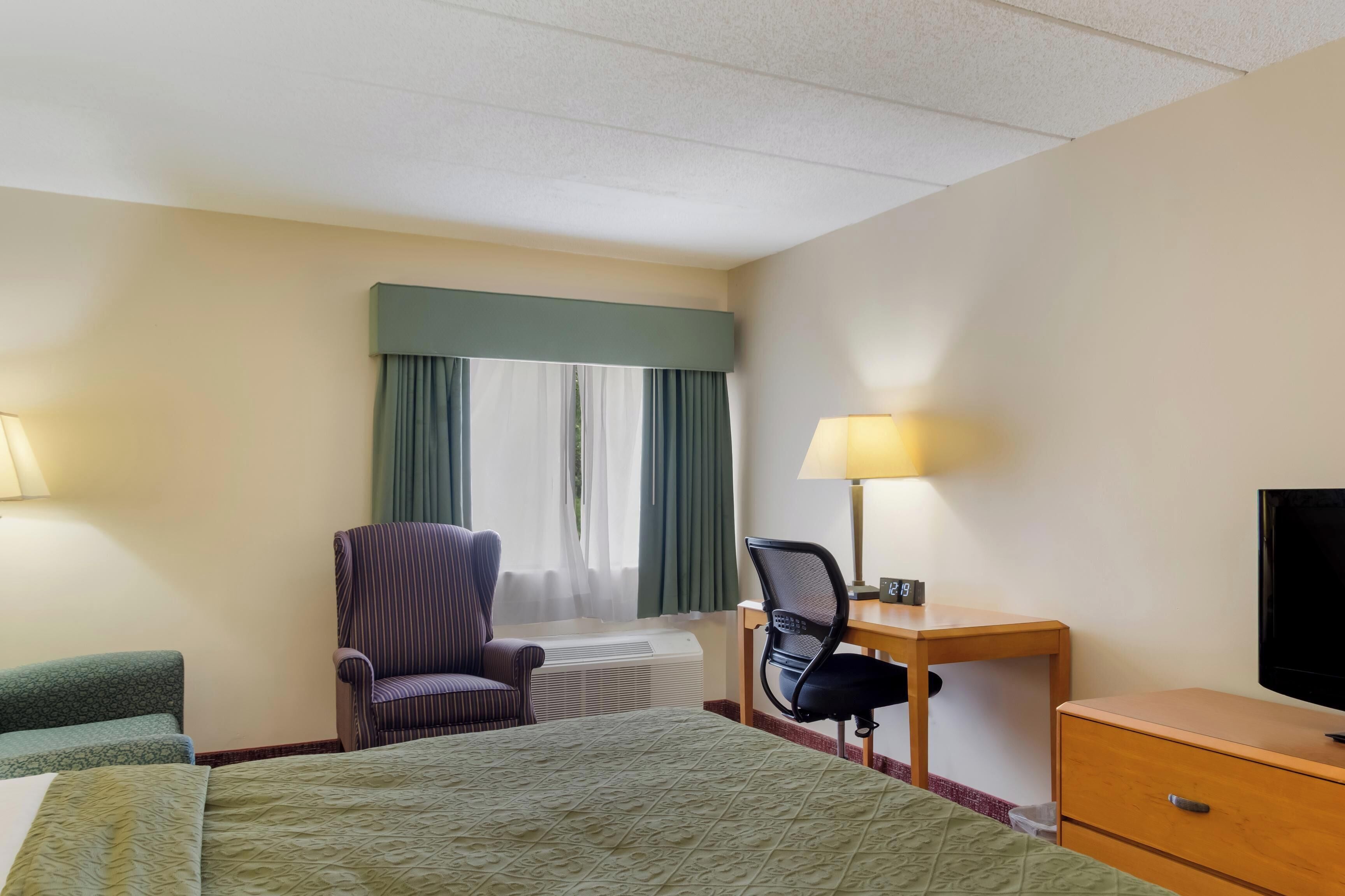 A hotel room with a bed, a desk with a chair, a television, and a window with curtains.