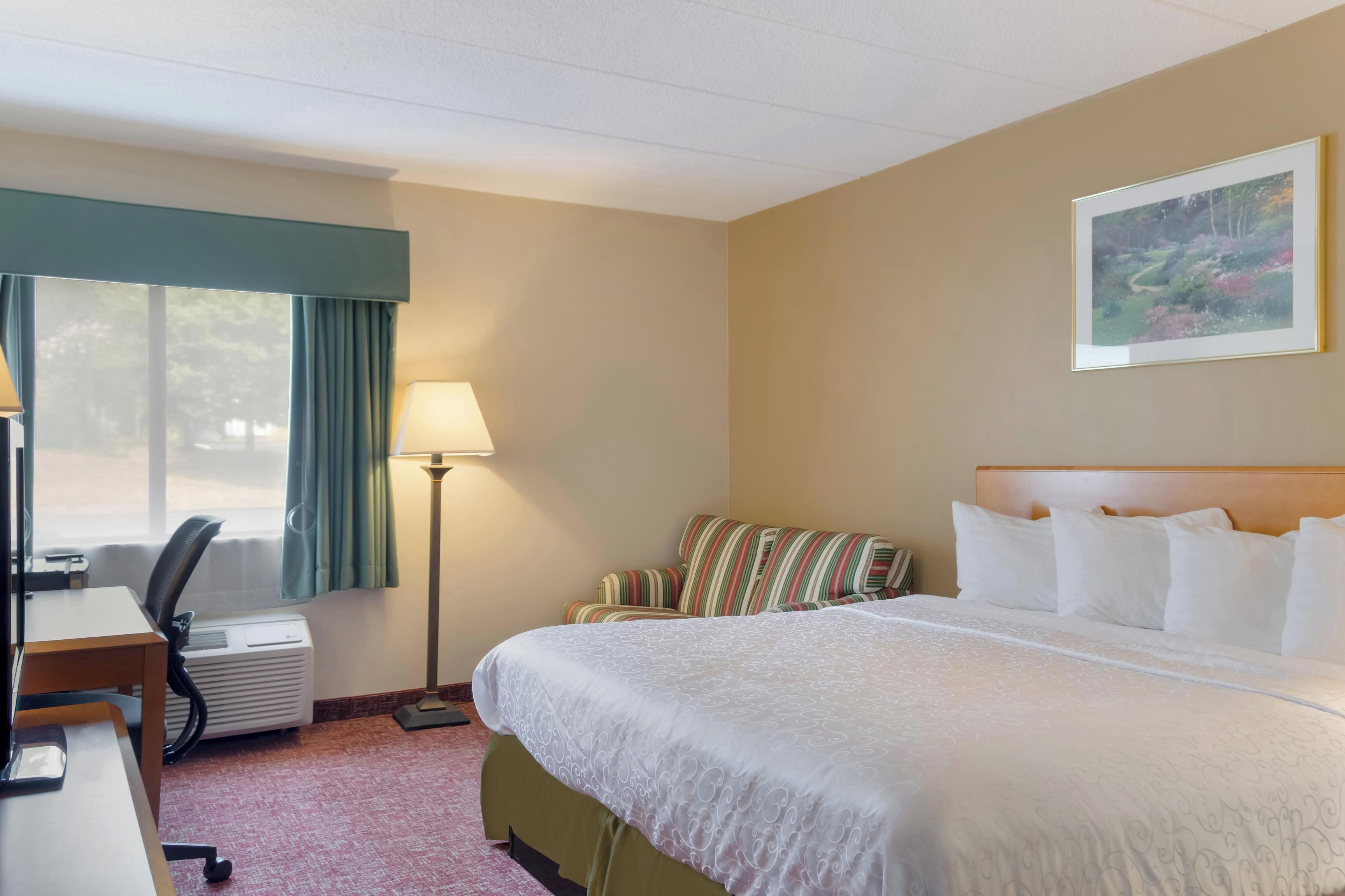 A hotel room with a large bed, a desk with a chair, a standing lamp, and a window with curtains.