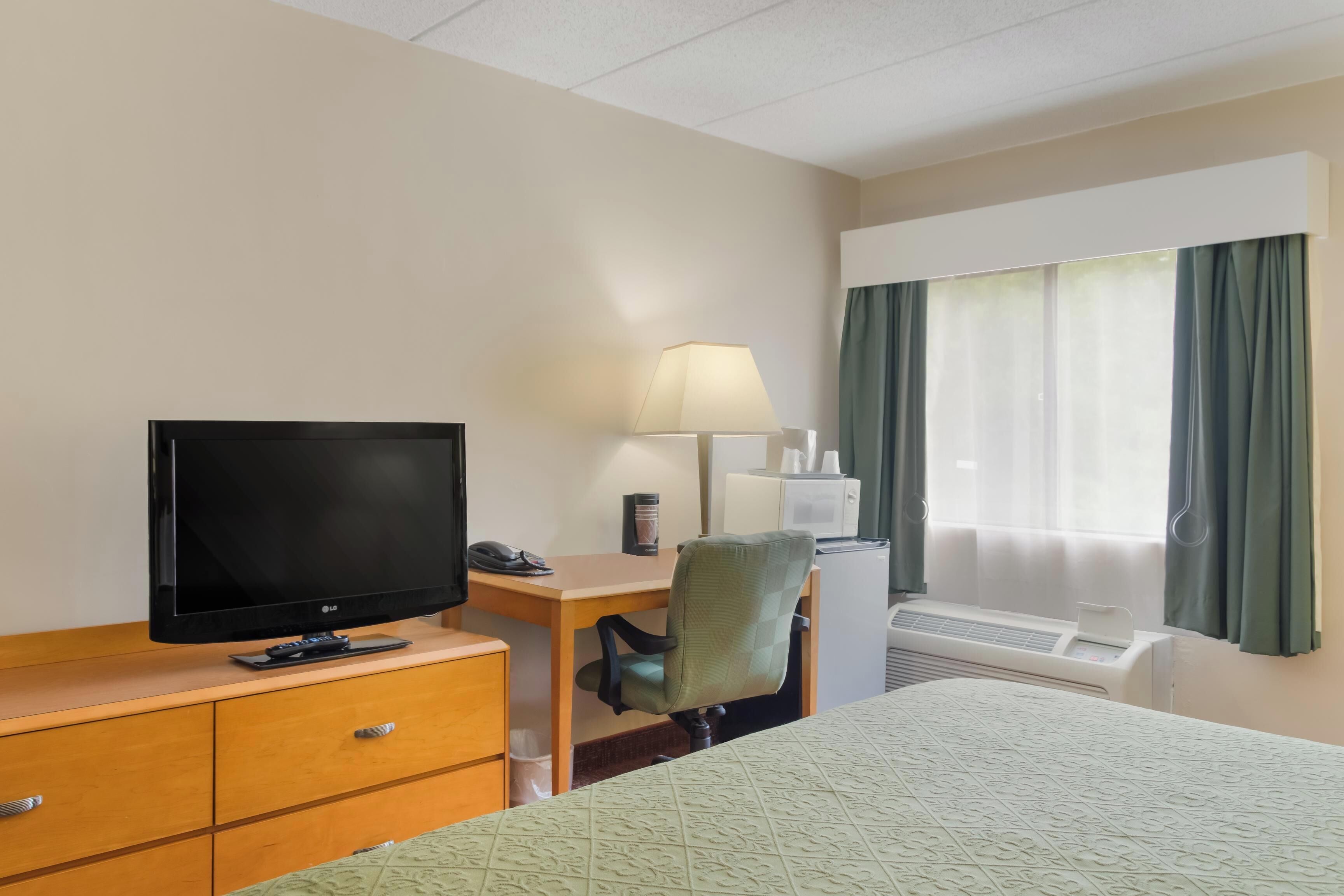 A hotel room with a bed, a desk with a lamp, a television, and an air conditioning unit.