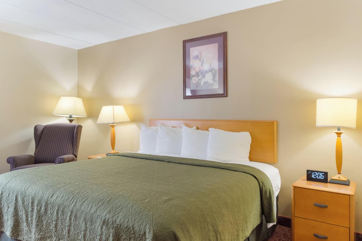 A hotel room with a bed, two bedside lamps, a chair, a nightstand with a clock, and a framed picture on the wall.