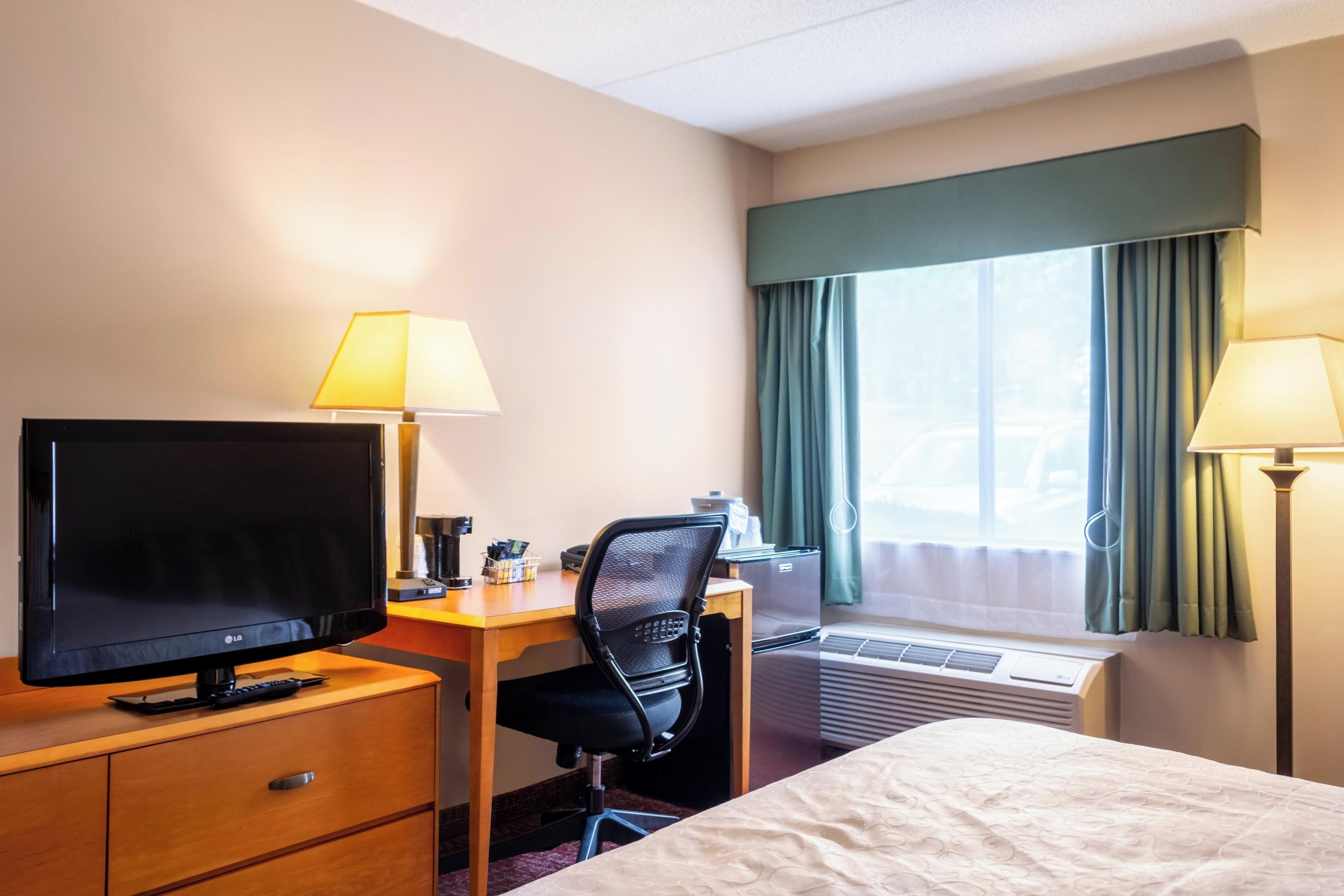 A hotel room with a bed, desk, TV, and air conditioning unit.
