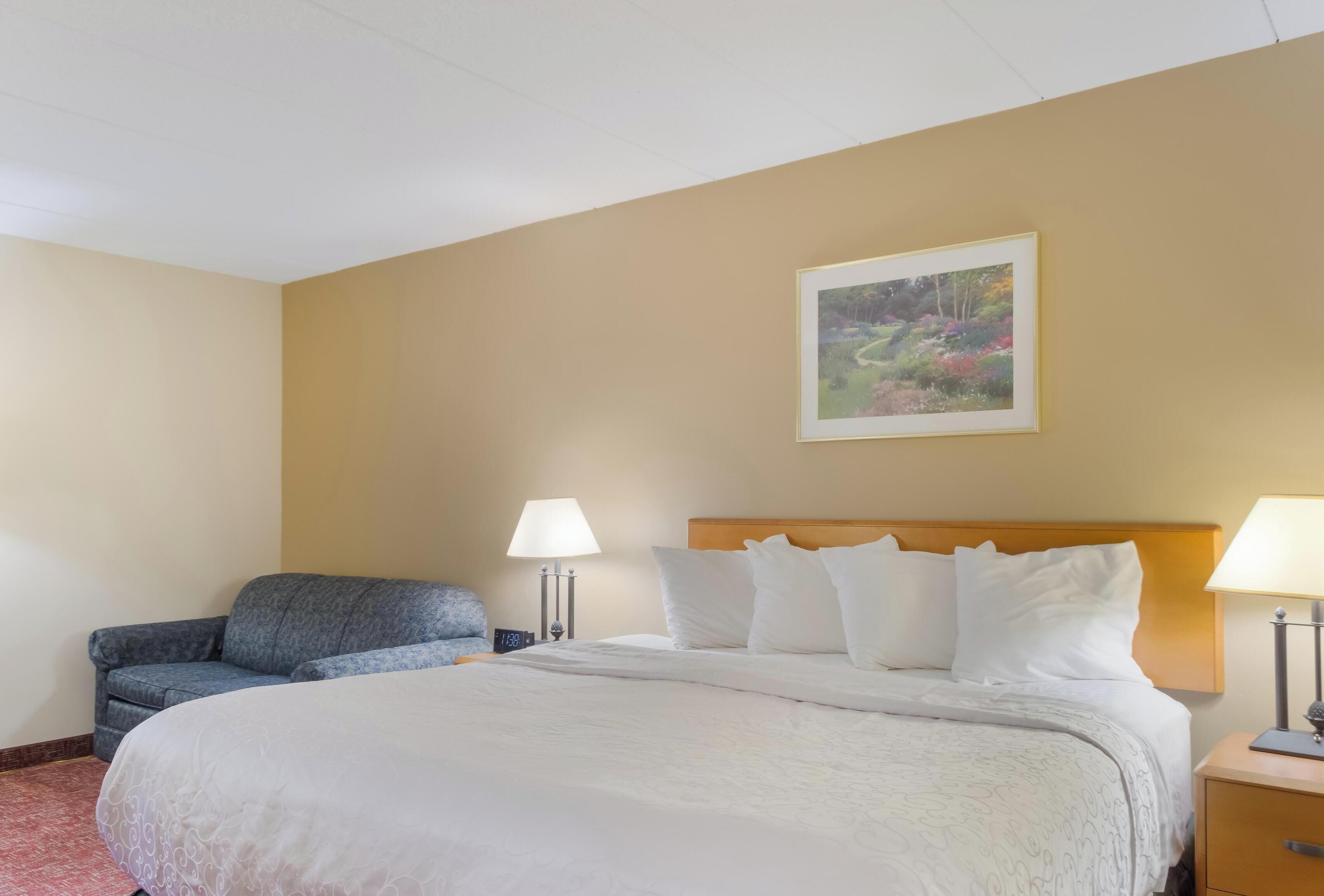 A hotel room with a large bed, two bedside lamps, a gray sofa, and a framed picture on the wall.