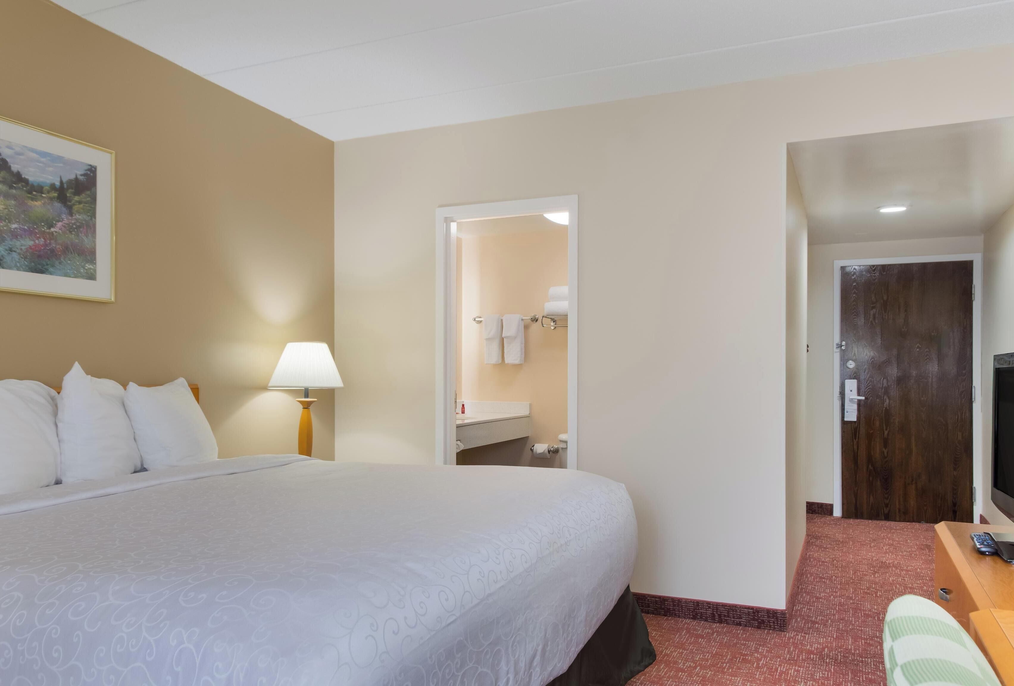 A hotel room with a large bed, a bedside lamp, a painting on the wall, a bathroom visible through an open door, and a desk with a chair.