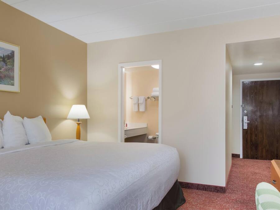A hotel room with a large bed, a bedside lamp, a painting on the wall, a bathroom visible through an open door, and a desk with a chair.