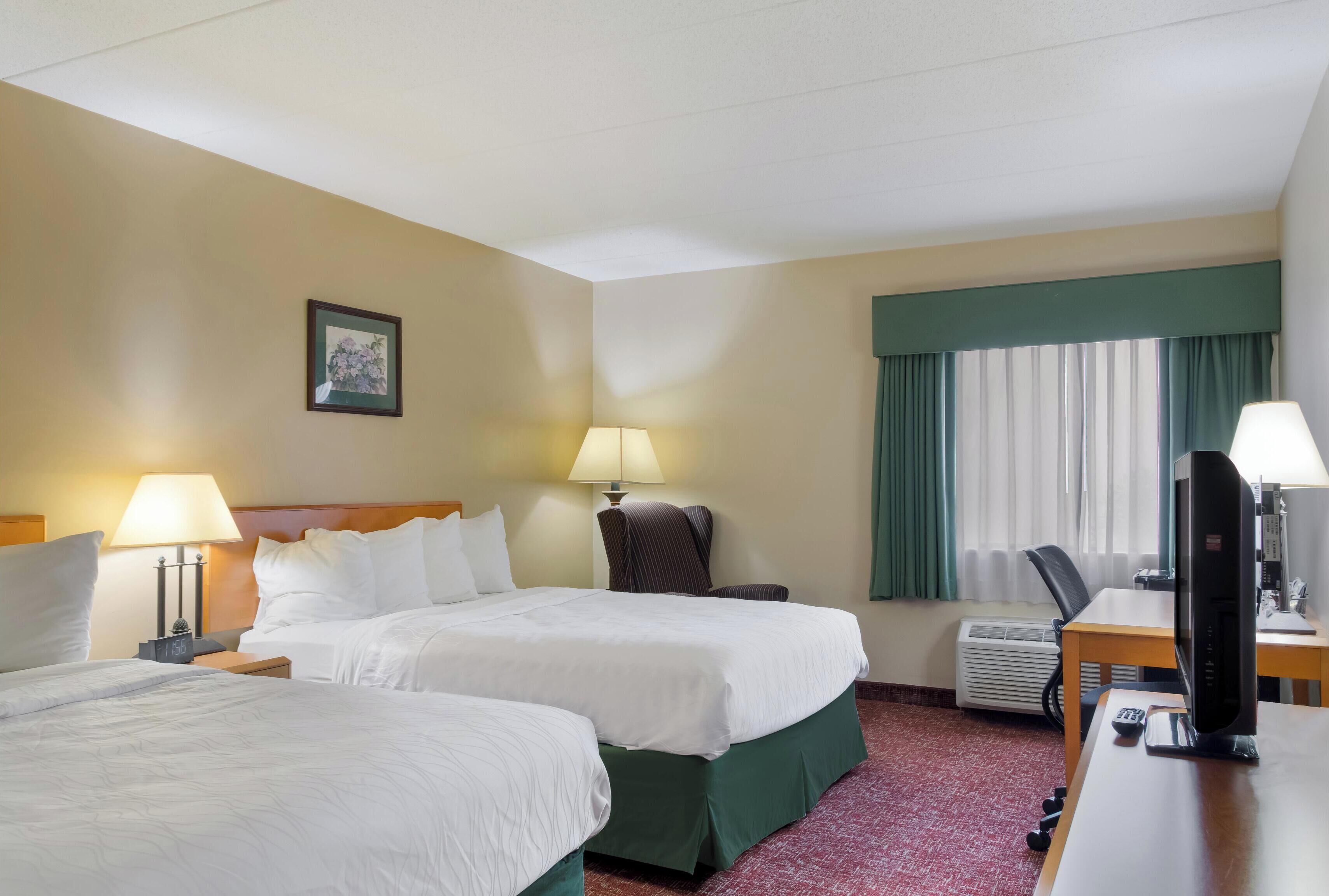 Een hotelkamer met twee bedden, een bureau, een computer, een stoel, een airconditioner en een raam met gordijnen.