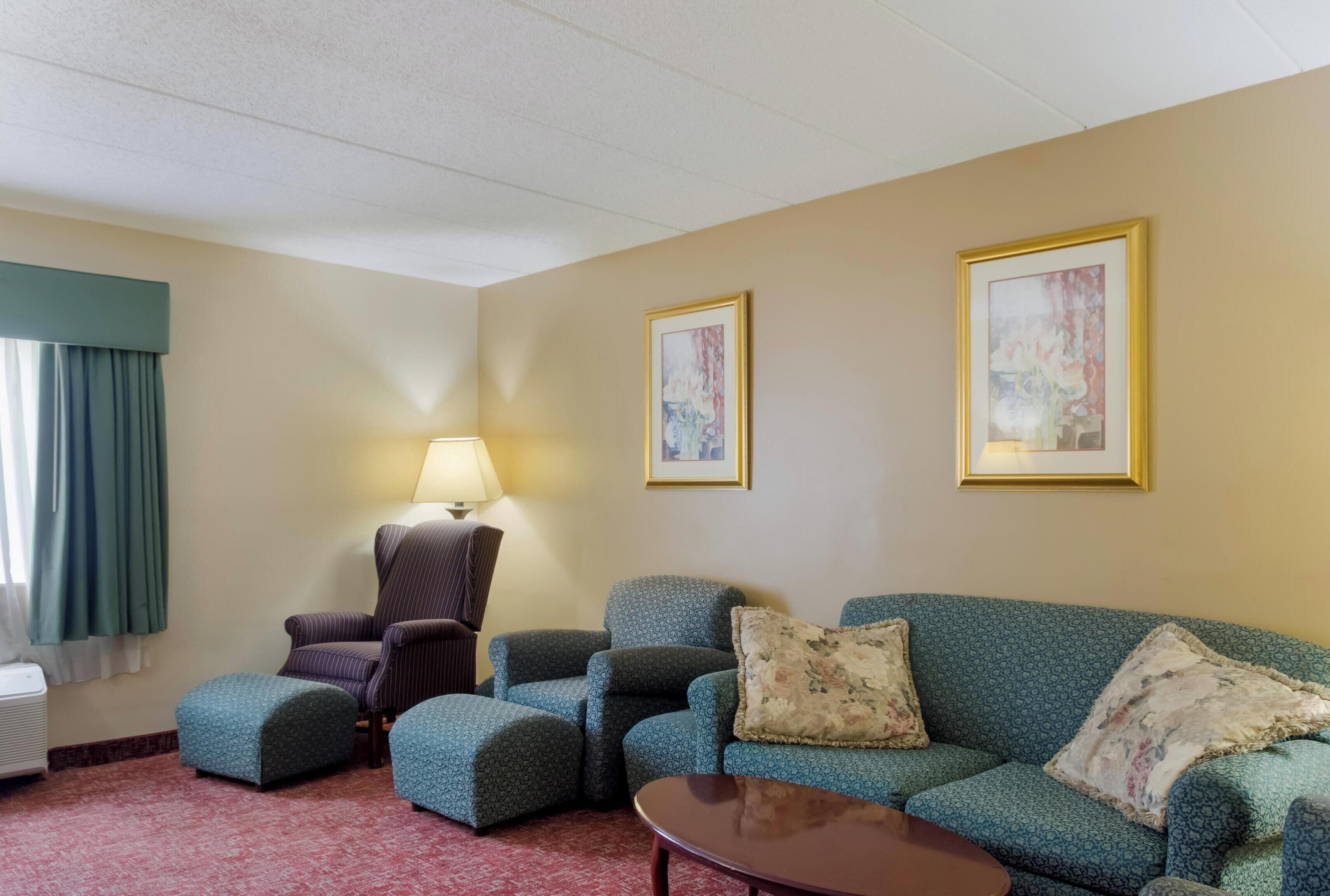 A hotel room with a green patterned sofa, two matching ottomans, a wooden coffee table, a striped armchair, a standing lamp, and two framed abstract paintings on the wall.