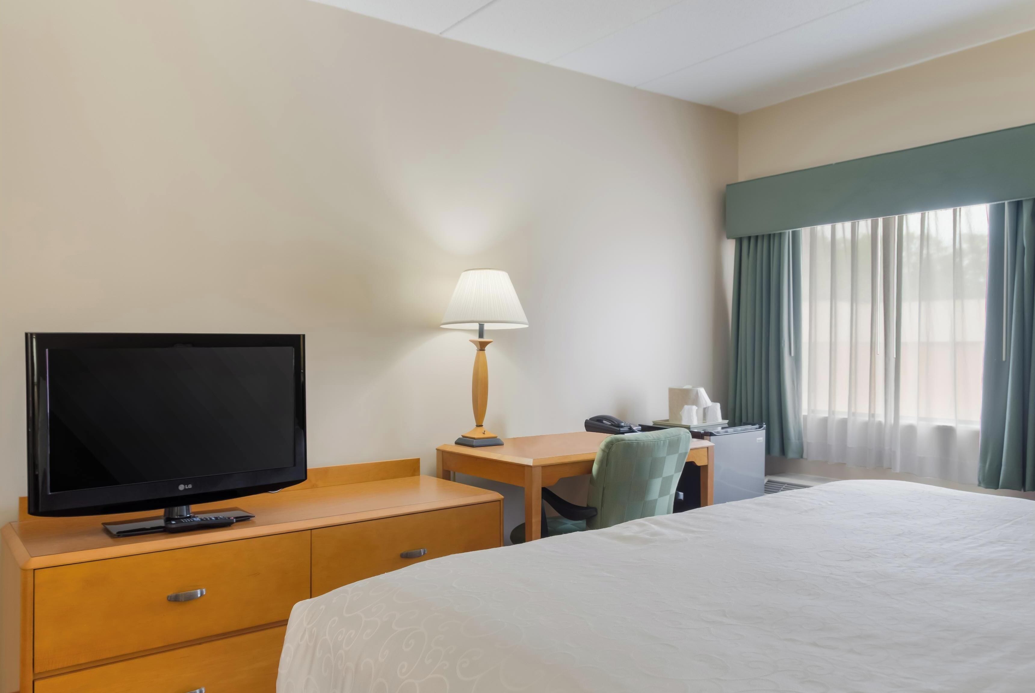 A hotel room with a bed, a TV, a desk with a lamp, a chair, and a window with curtains.