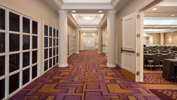 Un couloir d’hôtel avec un tapis à motifs, des colonnes blanches et un espace dédié aux conférences, comprenant des tables et des chaises.