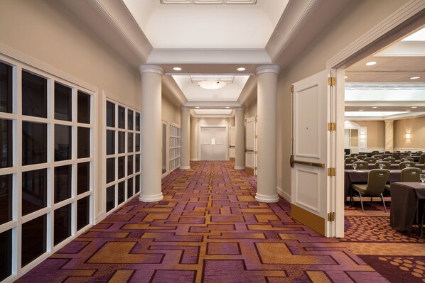 Un couloir d’hôtel avec un tapis à motifs, des colonnes blanches et un espace dédié aux conférences, comprenant des tables et des chaises.