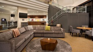 Een moderne hotellobby met een ruime hoekbank, een ronde salontafel en een trap naar een verdieping.