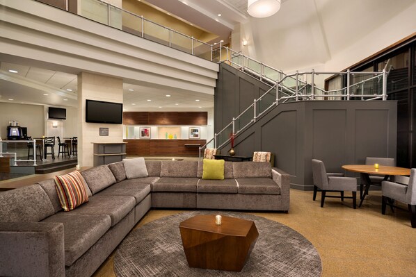 Un lobby moderno de hotel con un amplio sofá modular, una mesa de centro redonda y una escalera que conduce a un nivel superior.
