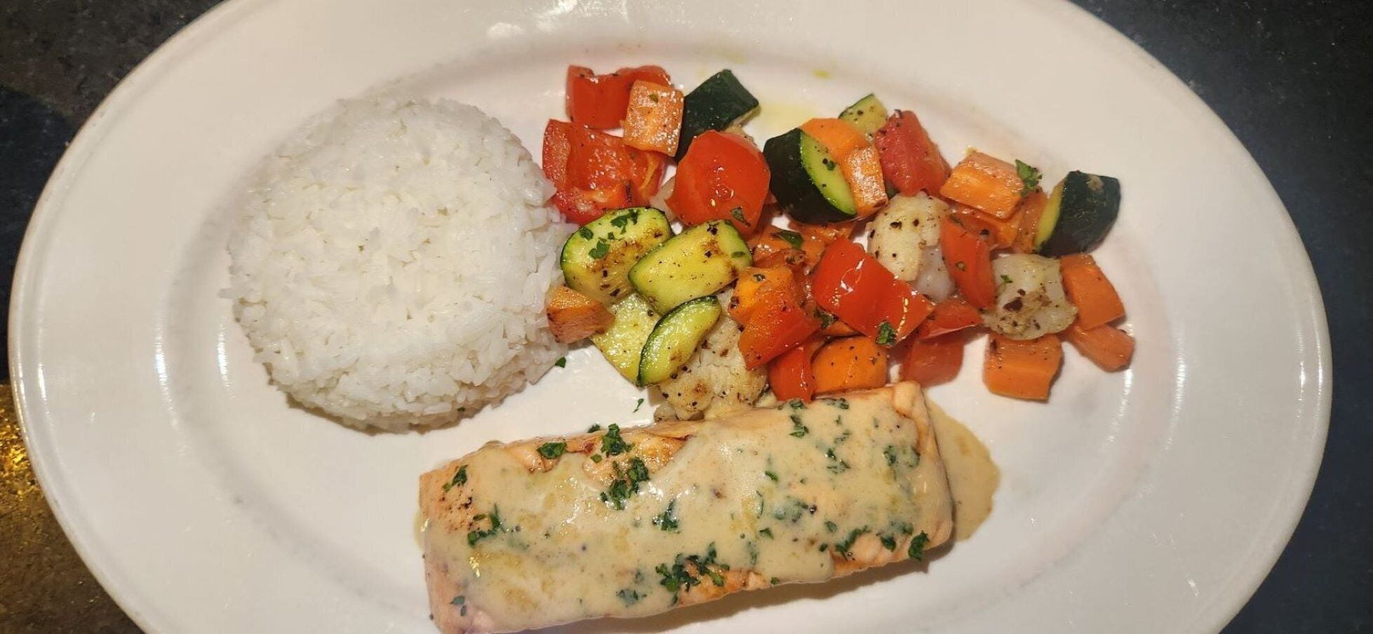 Ein Teller mit weißem Reis, ein Stück Lachs mit cremiger Soße und dazu ein Beilagensalat mit Karotten und Zucchini.