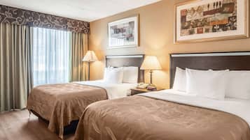 A hotel room with two beds, a large window with curtains, a framed picture on the wall, and a bedside table with a lamp.