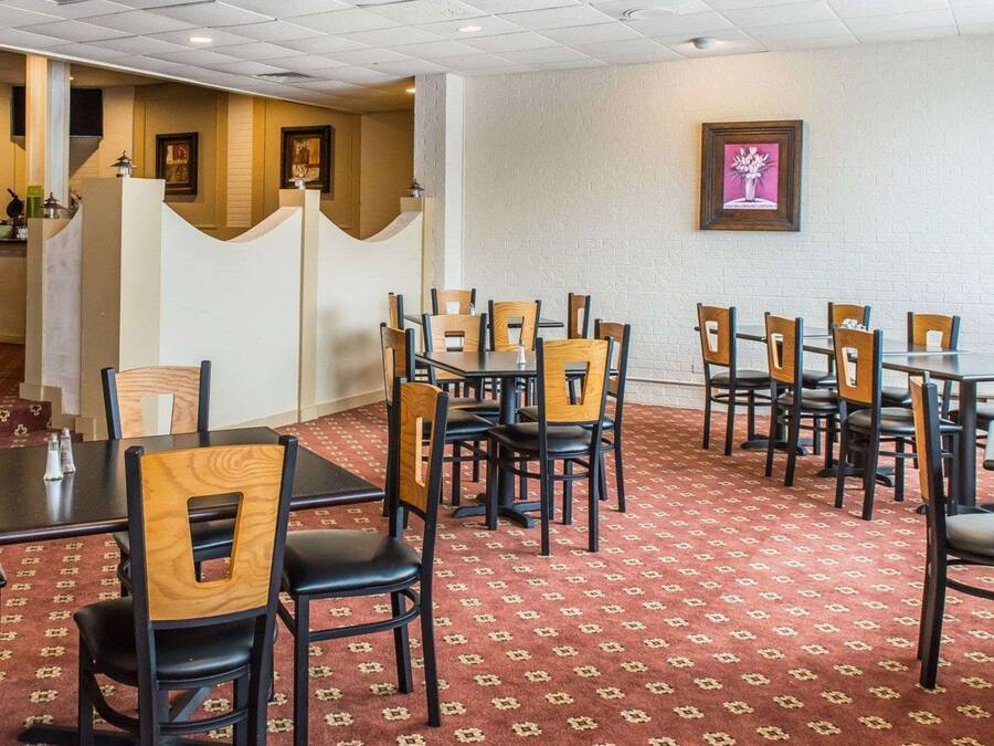 A hotel dining area with multiple tables and chairs, a patterned carpet, and a framed picture on the wall.