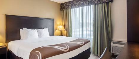 A hotel room with a large bed, a desk, a chair, a lamp, and a window with curtains.
