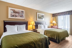 A hotel room with two beds, each with a green bedspread, a wooden headboard, and white bedding. There are two bedside tables with lamps, a framed picture on the wall, and a window with curtains.