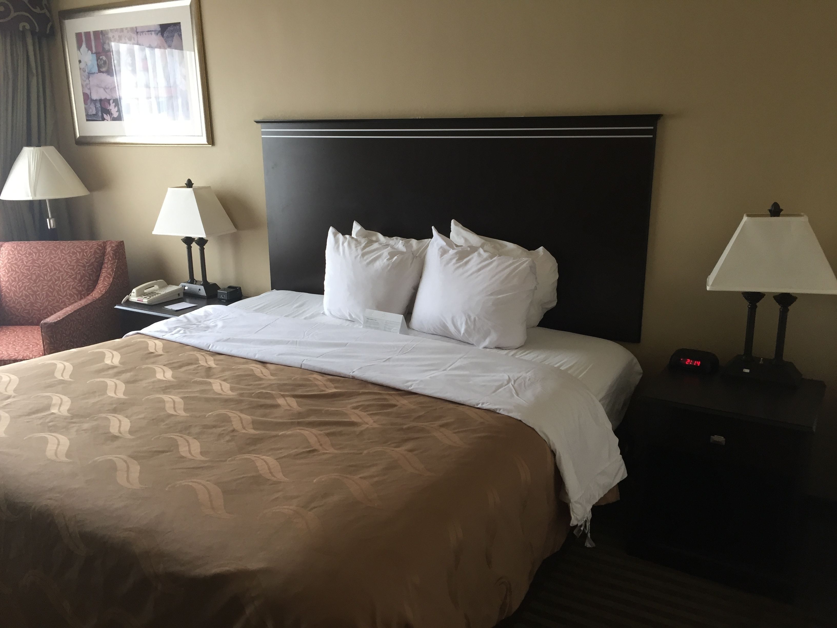 Habitación de hotel con una cama grande, dos lámparas de noche, una mesita de noche con un teléfono y un reloj digital, además de un sillón estampado.