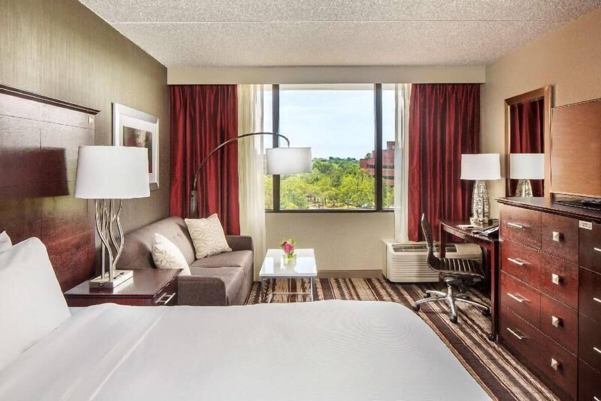 Habitación de hotel con cama, sofá, escritorio, cómoda y ventana con vistas a árboles y edificios.