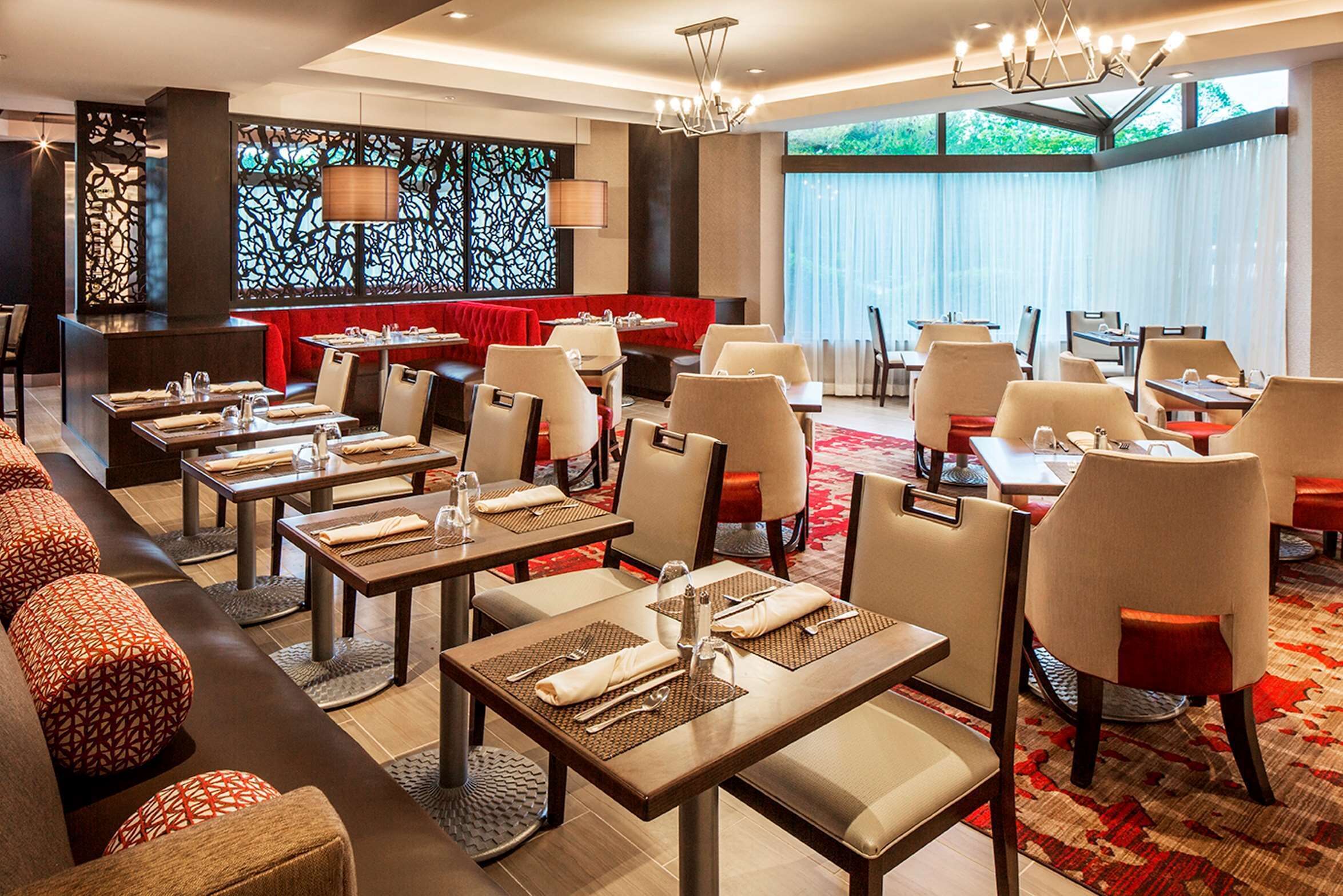 A modern restaurant with multiple dining tables and booths, a red cushioned seating area, and large windows providing natural light.