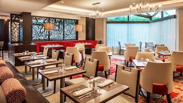 A modern restaurant with multiple dining tables and booths, a red cushioned seating area, and large windows providing natural light.