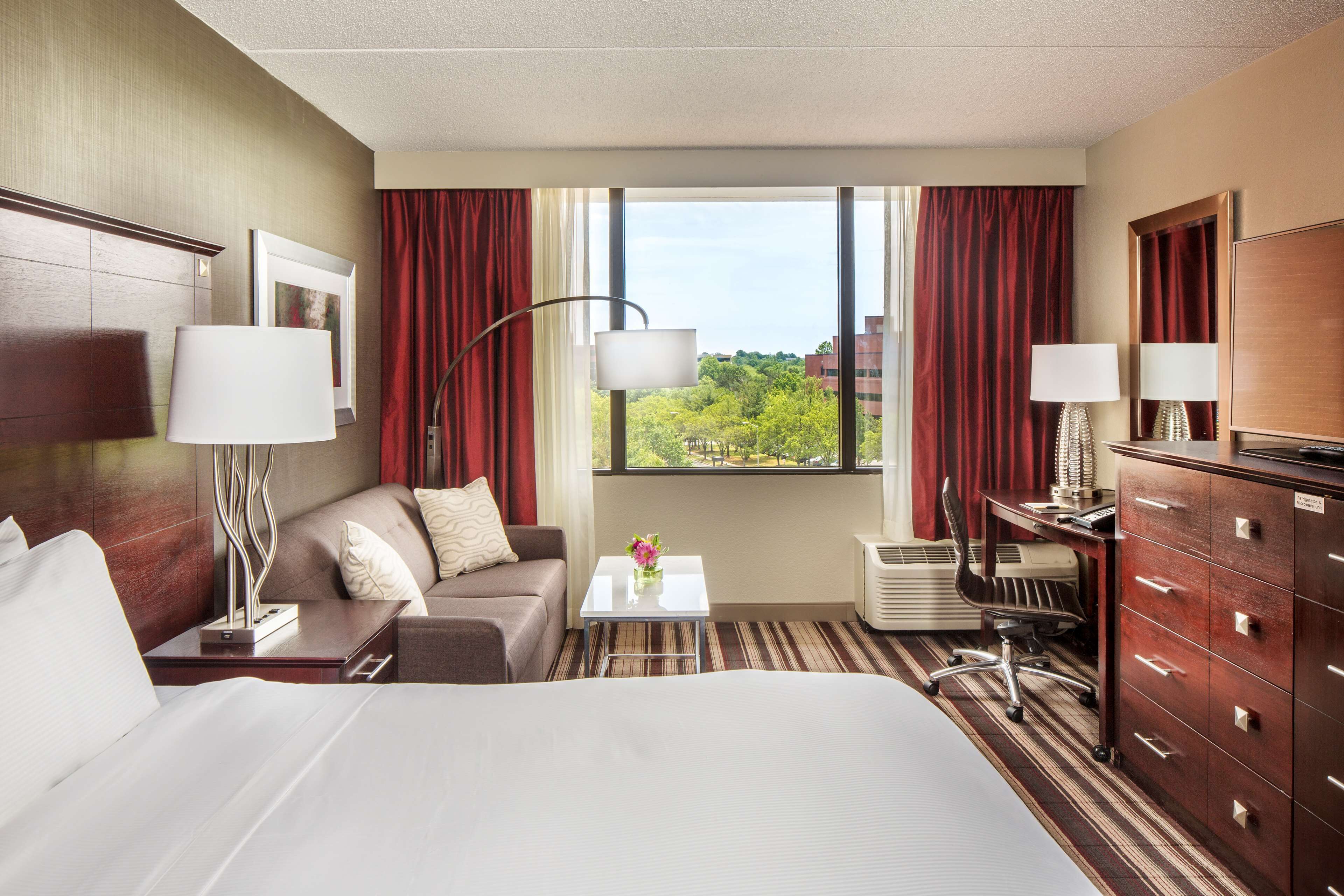 Quarto de hotel com uma cama grande, sofá, escrivaninha e cómoda. Há uma janela com vista para árvores e edifícios.
