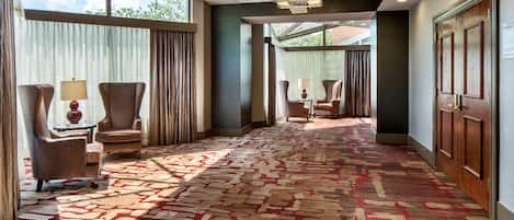 A hotel lobby with a patterned carpet, seating area with armchairs and lamps, and a large window with curtains.